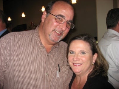 Man in beige shirt and lady in black top standing in dark room.