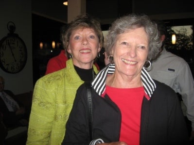 Lady in green coat and lady in black coat with red shirt standing in dark room.