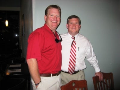 Man in red shirt and man in white shirt stanidng in front of white wall.