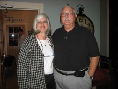Lady in black and white coat an man in black shirt standing in front of white wall with clock on wall.