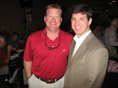 Man in red shirt and man in beige coat standing in room with dim light.