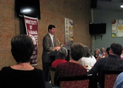 Man standing at podium with red banner in background.