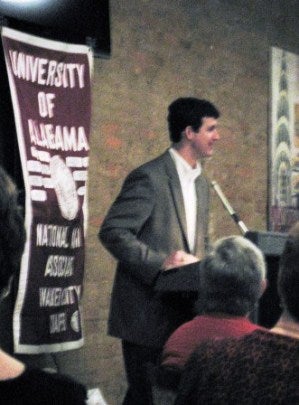 Man at Podium speaking with crimson banner in background.