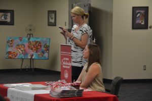 Lady sitting at table with red cloth and lady standing behind table.