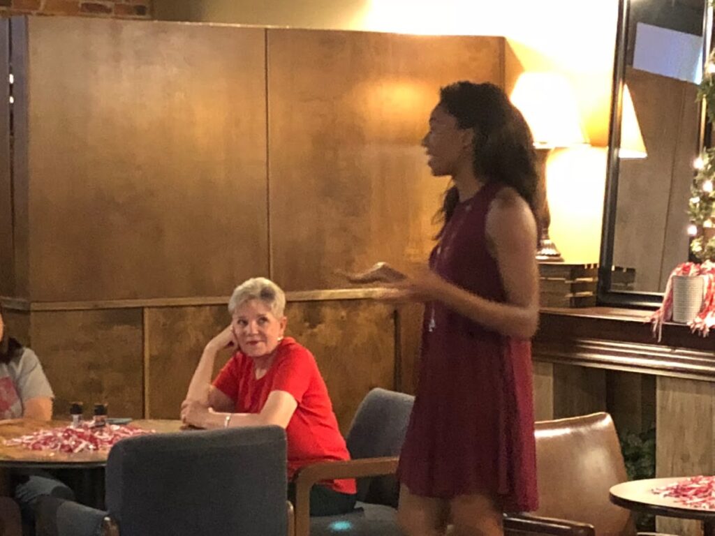 Two ladies one sitting in red shit and one standing in red dress in room with brown wall.