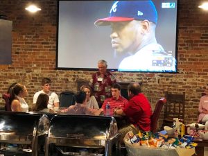 Group of people sitting in front of screen with picture of man in blue cap.