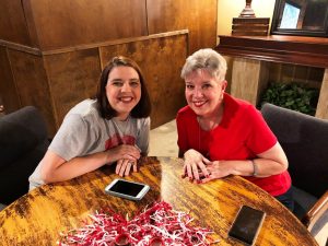 Two ladies sitting at wooden table with red and white shaker on table.