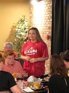 Group of people sitting at table and one lady in red shirt standing in front of brick wall.