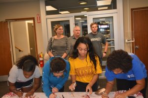 Group of people standing at table signing paper and group of people standing in background.