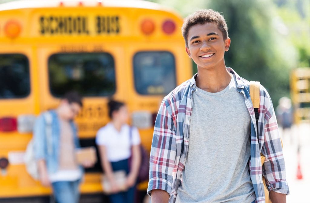A male high school student shown with a yellow school bus in the background